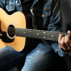 Un guitariste en jean montre comment tenir une guitare, ses doigts formant un accord sur le manche.