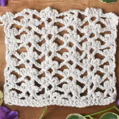 Échantillon de dentelle au crochet blanc, orné de motifs floraux délicats réalisés en triple crochet. Des feuilles vertes et des fleurs violettes entourent l'ouvrage.