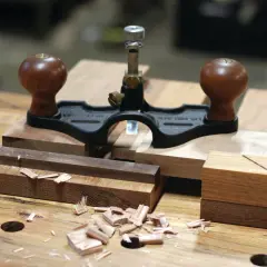 Une guimbarde, outil de menuisier, est posée sur des pièces de bois. Des copeaux témoignent du travail.