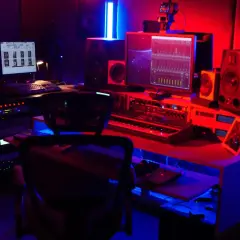 Studio d'enregistrement éclairé en rouge et bleu, avec un ordinateur affichant un logiciel de production musicale, un clavier, des enceintes et un sampler.