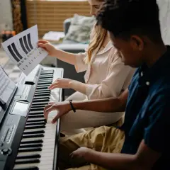 Deux mains jouent sur un piano, l'une suit une partition, l'autre une feuille avec des notes comme sol#m.