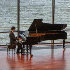 Un pianiste joue devant une grande fenêtre, sa pédale piano effleurant le sol. L'eau tourbillonnante à l'extérieur reflète la lumière.