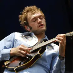 Un musicien aux yeux fermés joue de la mandoline, son visage exprimant la concentration. Sa chemise bleue est ornée de motifs colorés. La musique mandoline connue résonne.