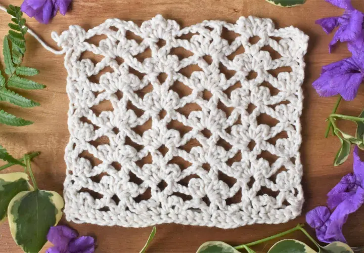 Échantillon de dentelle au crochet blanc, orné de motifs floraux délicats réalisés en triple crochet. Des feuilles vertes et des fleurs violettes entourent l'ouvrage.
