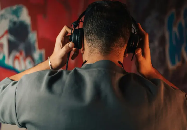 Un homme ajuste un type de casque audio sur ses oreilles, le dos tourné, devant un mur couvert de graffitis colorés.
