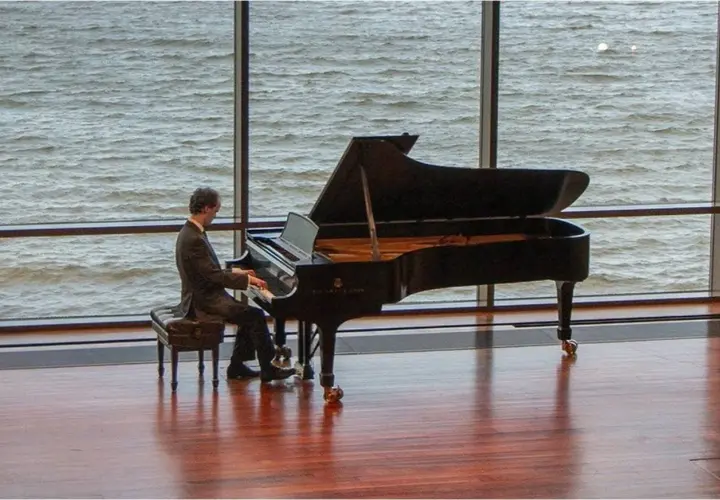 Un pianiste joue devant une grande fenêtre, sa pédale piano effleurant le sol. L'eau tourbillonnante à l'extérieur reflète la lumière.