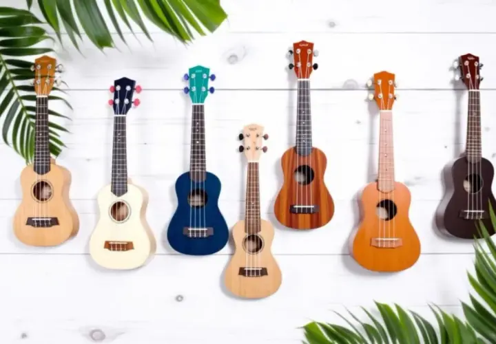 Sept ukulélés de différentes couleurs et tailles, dont un ukulélé de taille soprano, sont accrochés sur un mur en bois blanc, entourés de feuilles vertes.