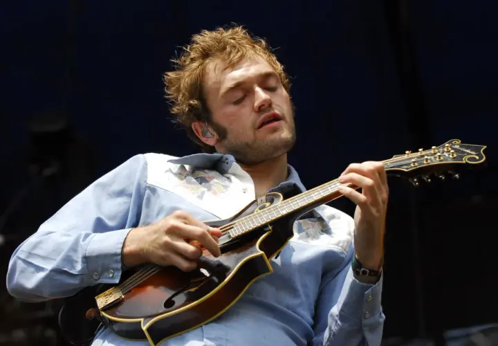Un musicien aux yeux fermés joue de la mandoline, son visage exprimant la concentration. Sa chemise bleue est ornée de motifs colorés. La musique mandoline connue résonne.