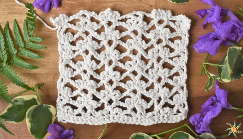Échantillon de dentelle au crochet blanc, orné de motifs floraux délicats réalisés en triple crochet. Des feuilles vertes et des fleurs violettes entourent l'ouvrage.