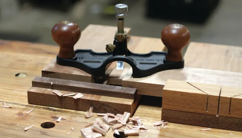 Une guimbarde, outil de menuisier, est posée sur des pièces de bois. Des copeaux témoignent du travail.