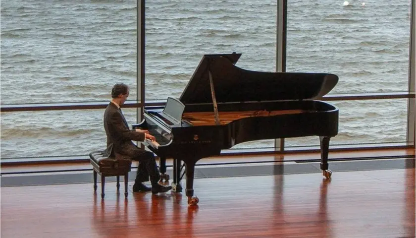 Un pianiste joue devant une grande fenêtre, sa pédale piano effleurant le sol. L'eau tourbillonnante à l'extérieur reflète la lumière.