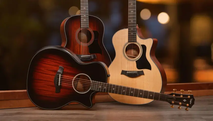 Trois guitares acoustiques, dont une en palissandre, posées sur un parquet en bois. L'arrière-plan est flou et lumineux.