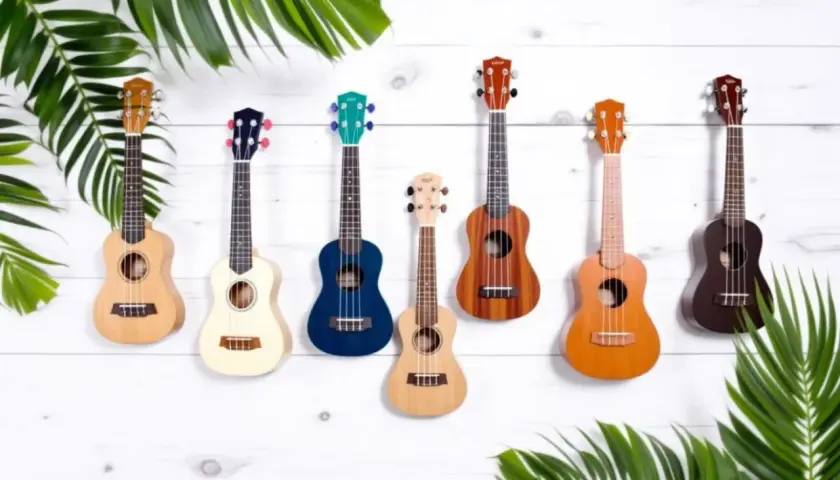 Sept ukulélés de différentes couleurs et tailles, dont un ukulélé de taille soprano, sont accrochés sur un mur en bois blanc, entourés de feuilles vertes.