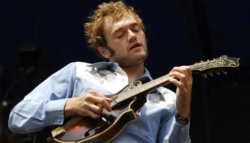 Un musicien aux yeux fermés joue de la mandoline, son visage exprimant la concentration. Sa chemise bleue est ornée de motifs colorés. La musique mandoline connue résonne.