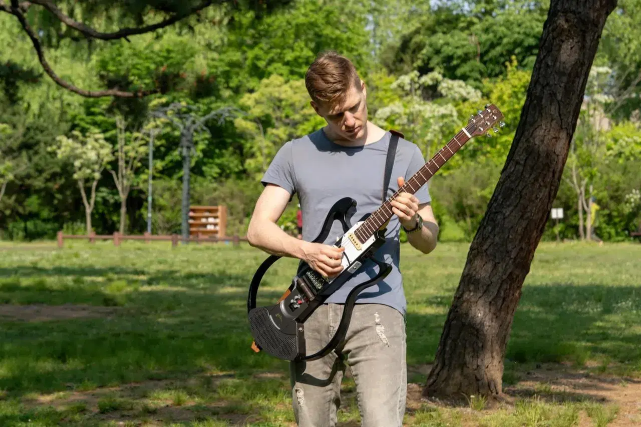 Un homme joue d'une guitare électrique sans ampli dans un parc verdoyant, profitant du beau temps pour faire de la musique en plein air.