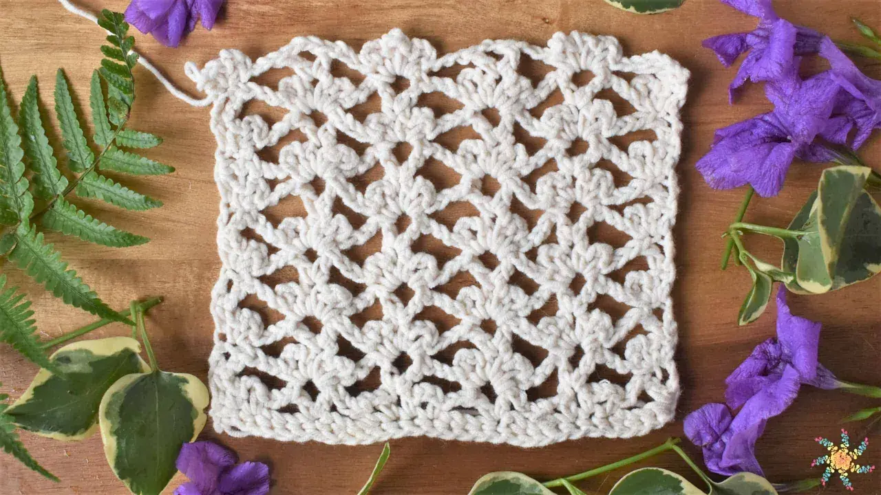 Échantillon de dentelle au crochet blanc, orné de motifs floraux délicats réalisés en triple crochet. Des feuilles vertes et des fleurs violettes entourent l'ouvrage.