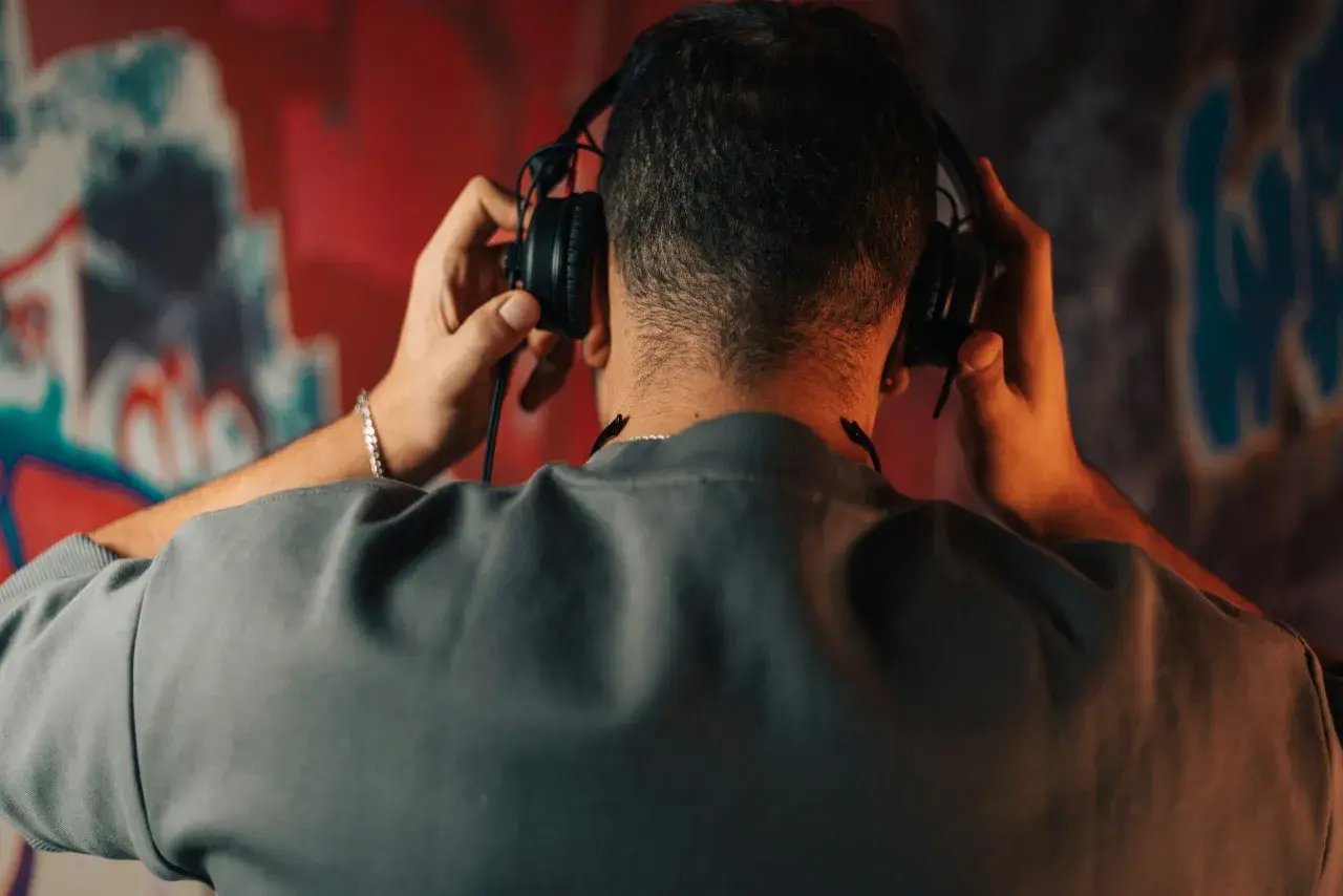 Un homme ajuste un type de casque audio sur ses oreilles, le dos tourné, devant un mur couvert de graffitis colorés.