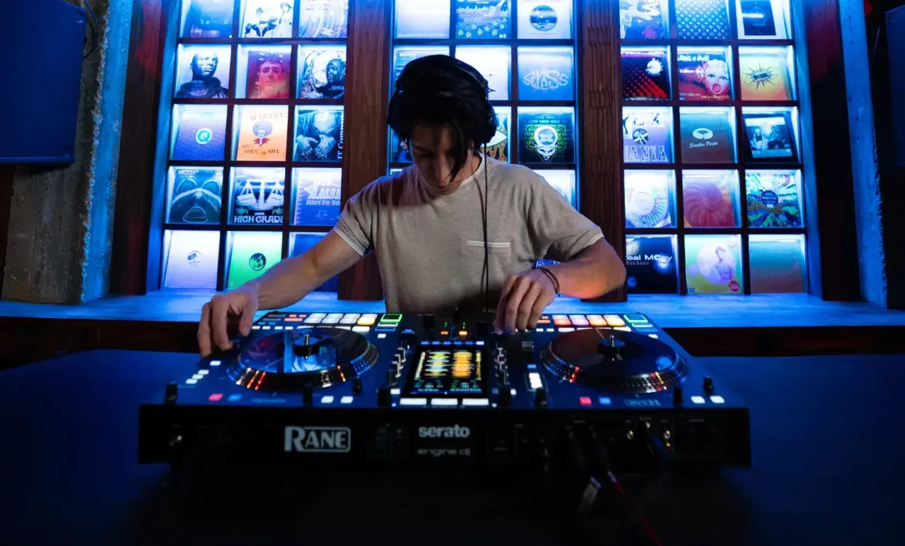 Un DJ mixe sur une console Rane, avec des vinyles rétro éclairés en fond. Fiche technique DJ.
