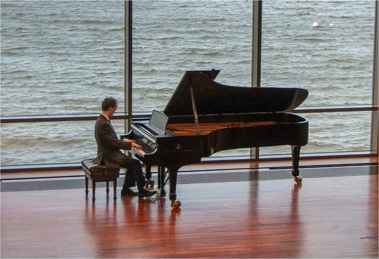 Un pianiste joue devant une grande fenêtre, sa pédale piano effleurant le sol. L'eau tourbillonnante à l'extérieur reflète la lumière.