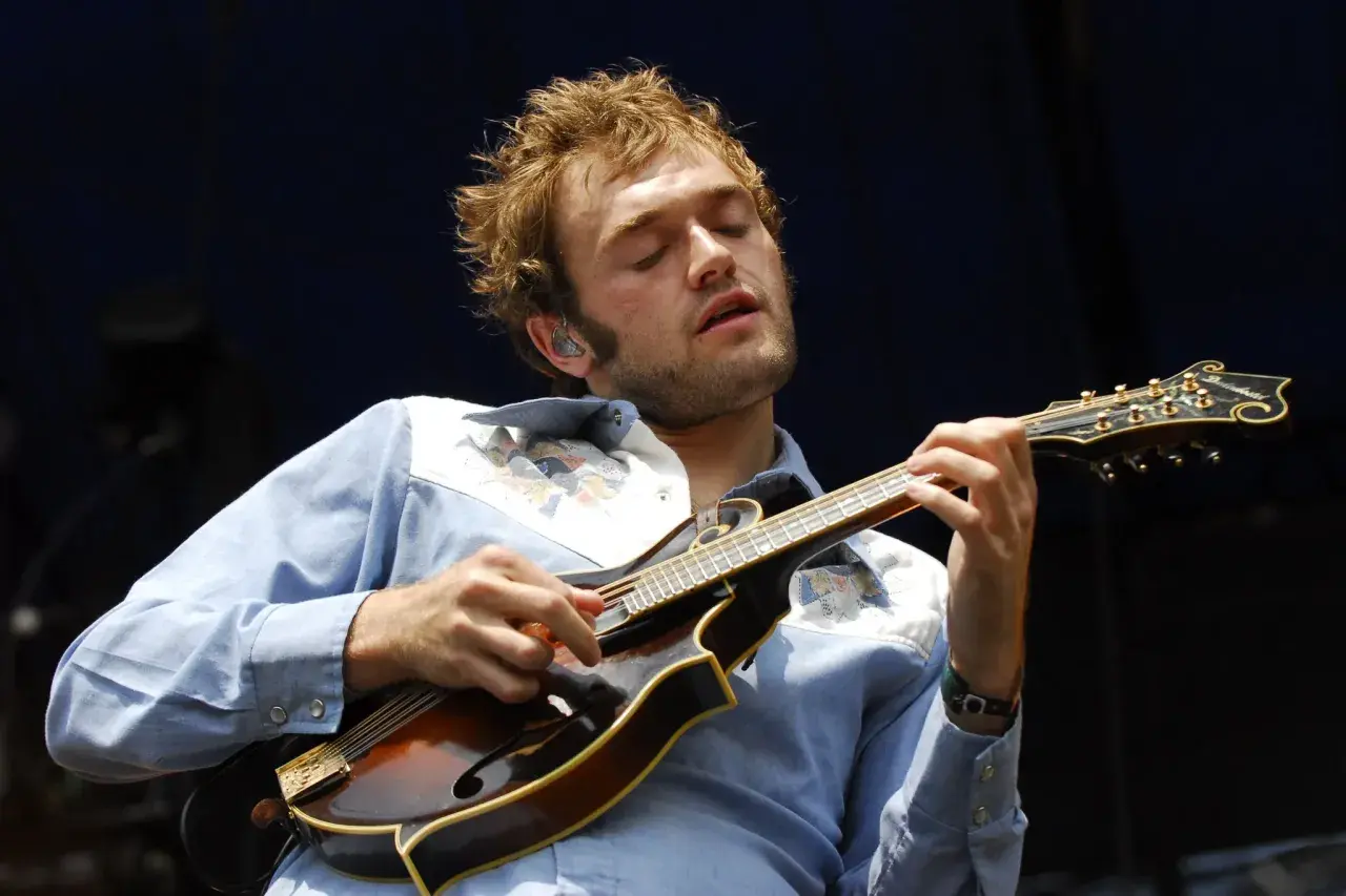 Un musicien aux yeux fermés joue de la mandoline, son visage exprimant la concentration. Sa chemise bleue est ornée de motifs colorés. La musique mandoline connue résonne.