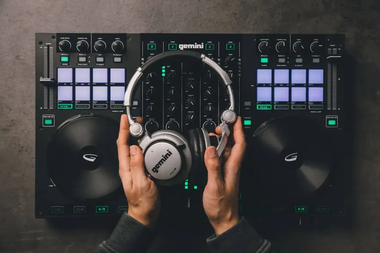 Mains d'un DJ tenant un casque argenté au-dessus d'une console de mixage Gemini. Le setup DJ est prêt pour la musique.