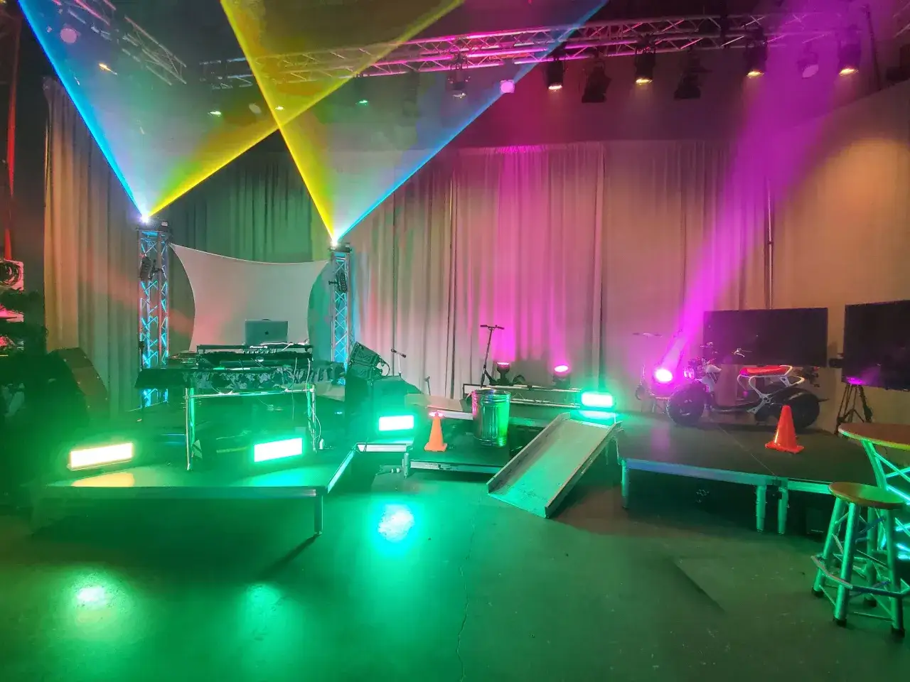 Scène prête pour un enregistrement live : DJ, lumières colorées, trottinettes et un scooter. L'ambiance est électrique !