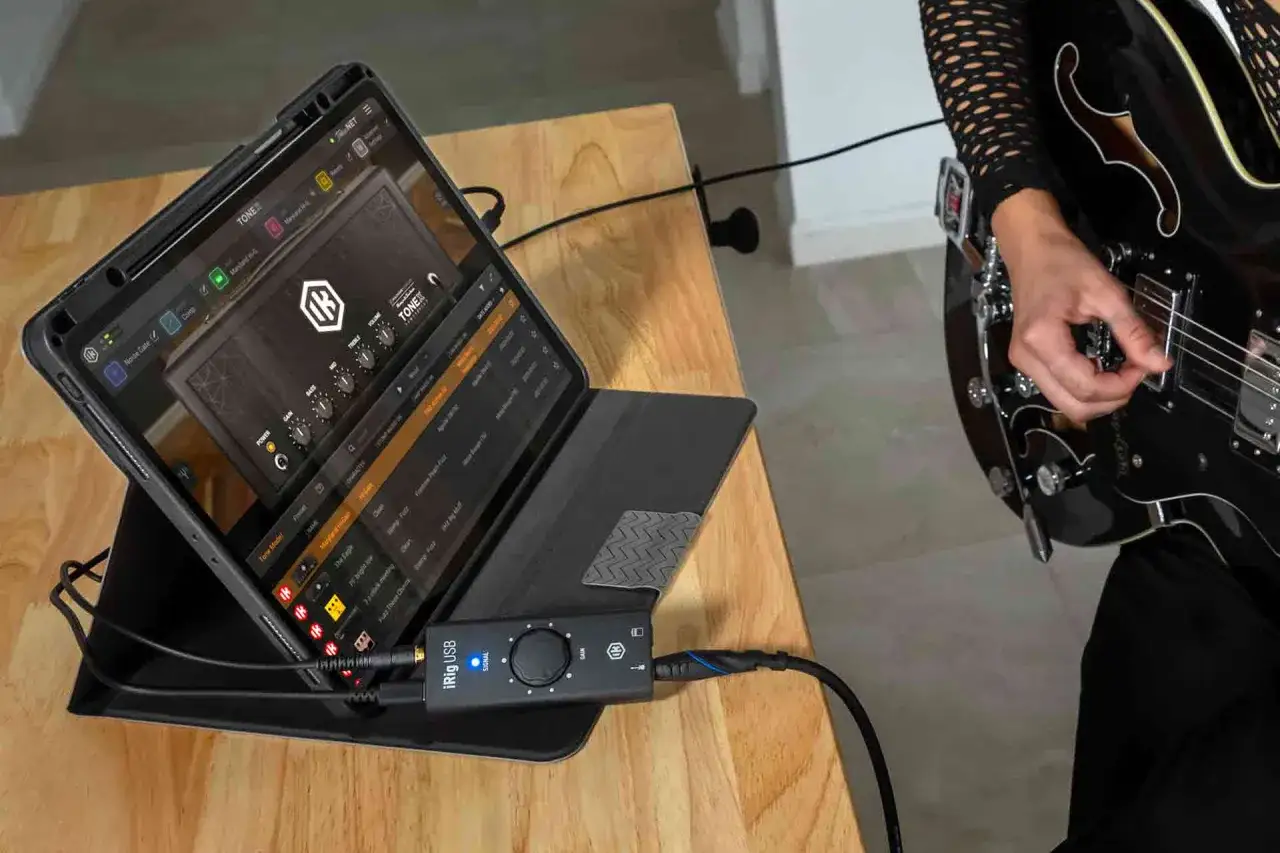 Une personne joue de la guitare, connectée à une tablette via un adaptateur iRig USB pour brancher guitare sur PC et explorer des sons virtuels.