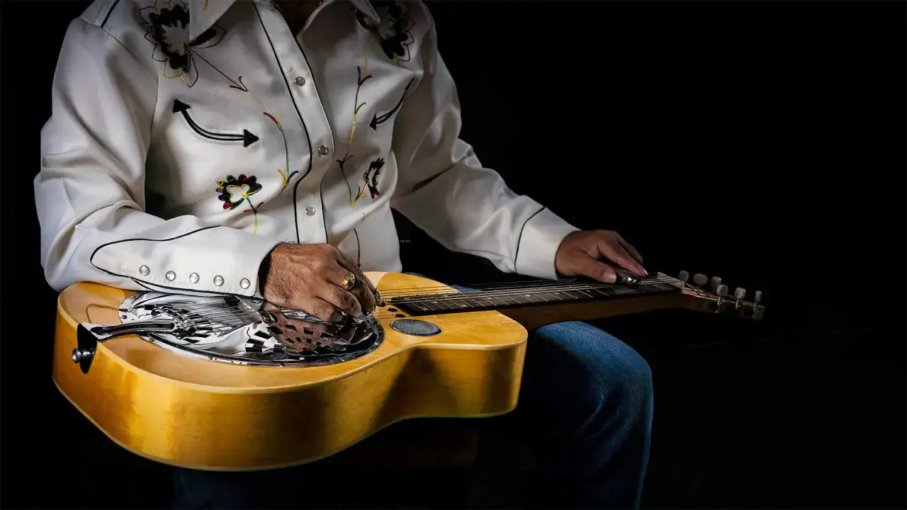 Un musicien joue une guitare dobro blues, sa chemise brod&eacute;e contrastant avec le bois clair de l'instrument.