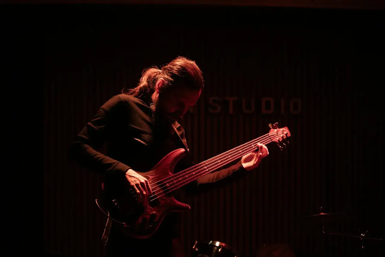 Un musicien aux cheveux longs, concentr&eacute;, joue un solo de basse &eacute;lectrique dans une ambiance tamis&eacute;e.