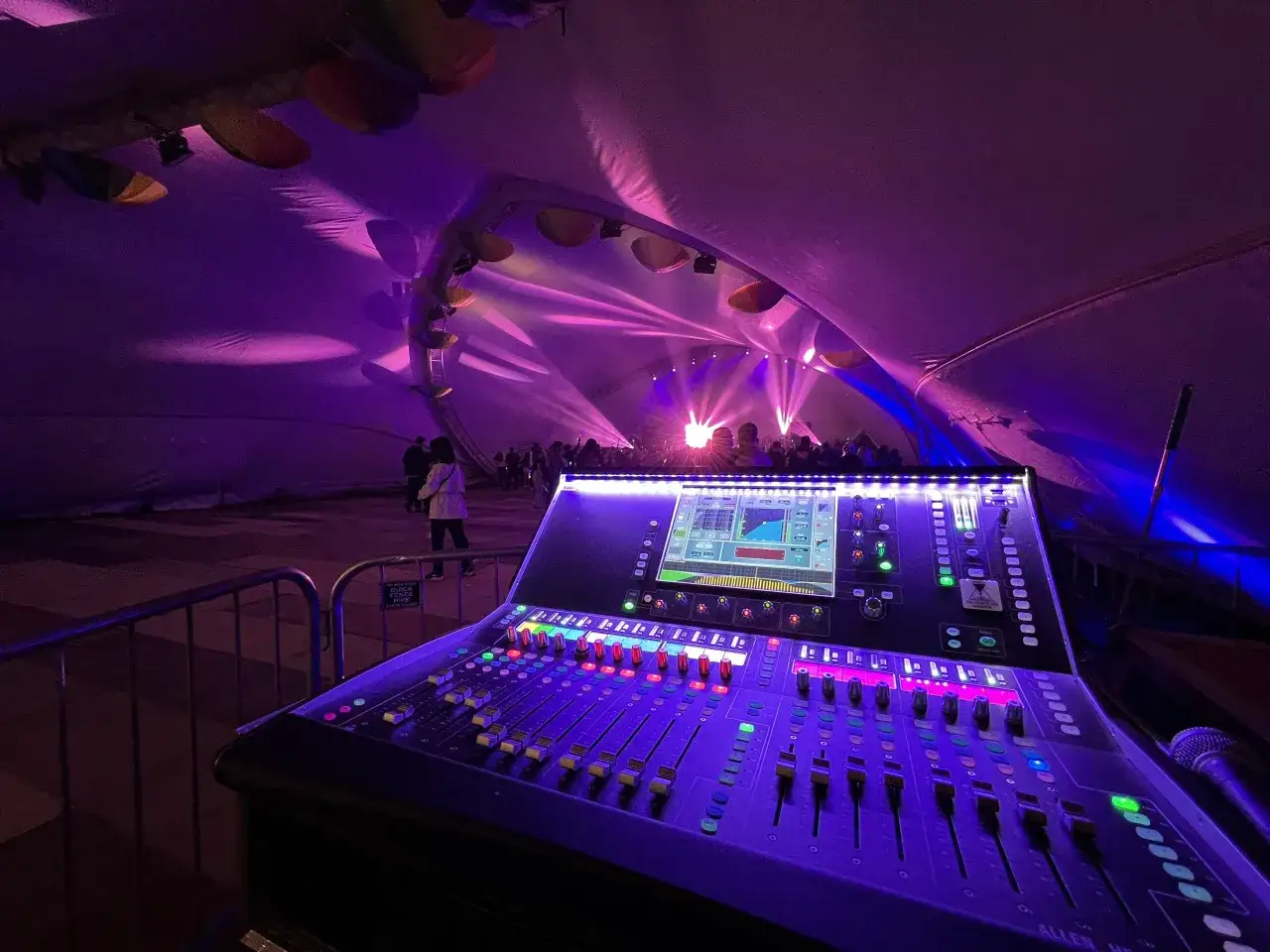 Console de mixage illumin&eacute;e, pr&ecirc;te pour le sound check. Lumi&egrave;res violettes et roses dans une tente, public au loin.