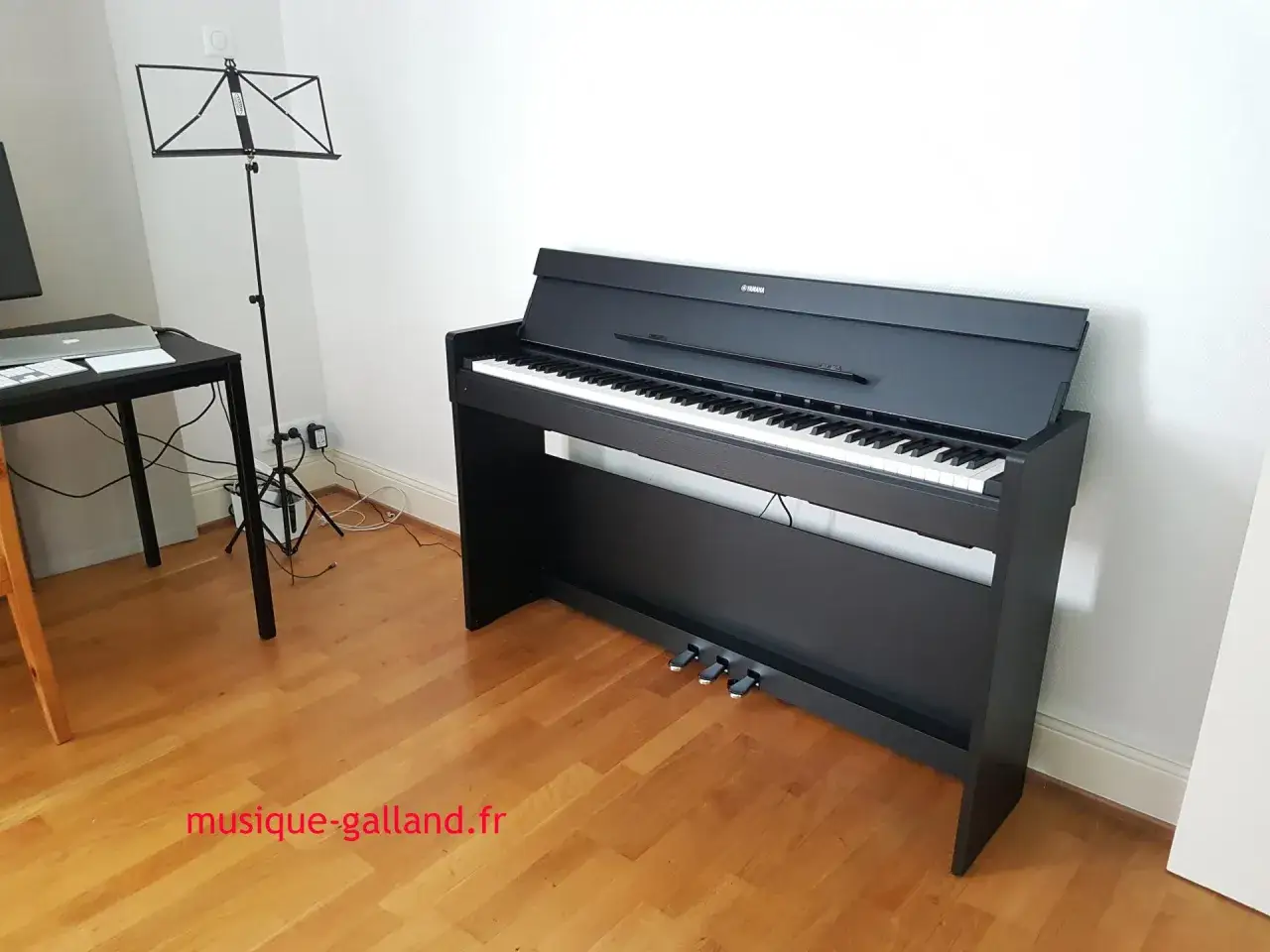 Un piano numérique Yamaha noir, prêt à jouer, avec un pupitre et un bureau à proximité.