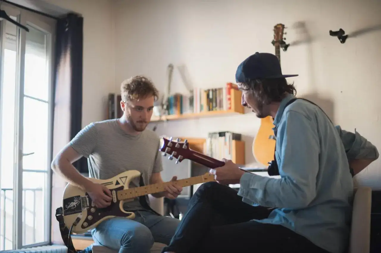 Deux hommes apprennent comment tenir une guitare, l'un jouant d'une guitare blanche, l'autre d'une guitare rouge.